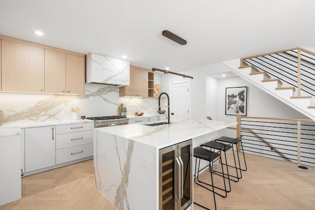 kitchen featuring wine cooler, light stone counters, a sink, light brown cabinetry, and an island with sink