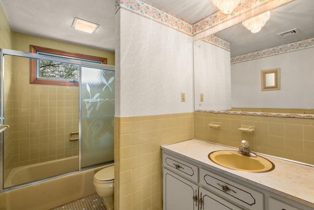 full bathroom with tile walls, toilet, bath / shower combo with glass door, vanity, and wainscoting