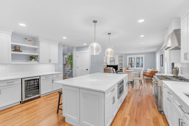 kitchen featuring wine cooler, appliances with stainless steel finishes, white cabinetry, light wood-type flooring, and wall chimney exhaust hood