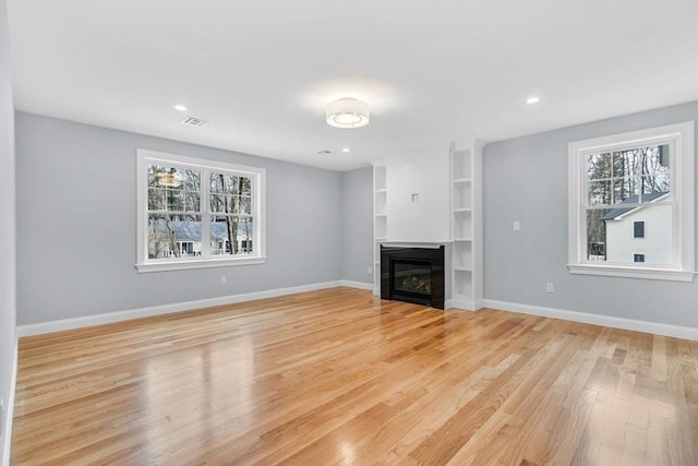 unfurnished living room with built in shelves, visible vents, baseboards, light wood-type flooring, and a glass covered fireplace