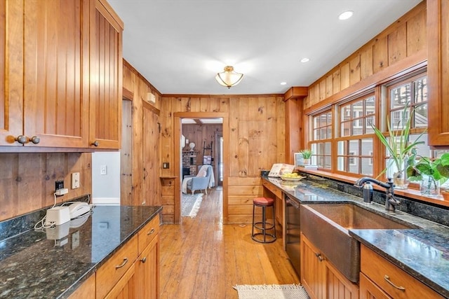 kitchen featuring dark stone counters, dishwashing machine, light wood-style flooring, wood walls, and a sink