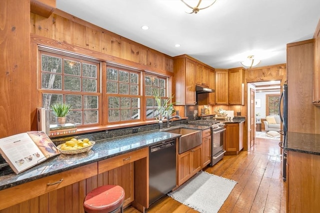 kitchen with stainless steel appliances, brown cabinetry, a sink, dark stone counters, and under cabinet range hood