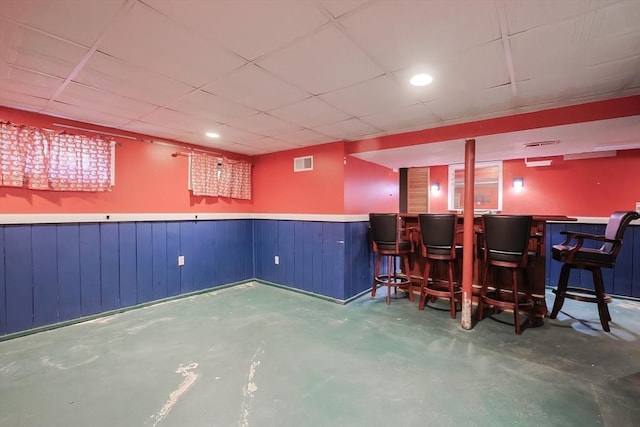 bar with visible vents, a paneled ceiling, wainscoting, a bar, and concrete floors