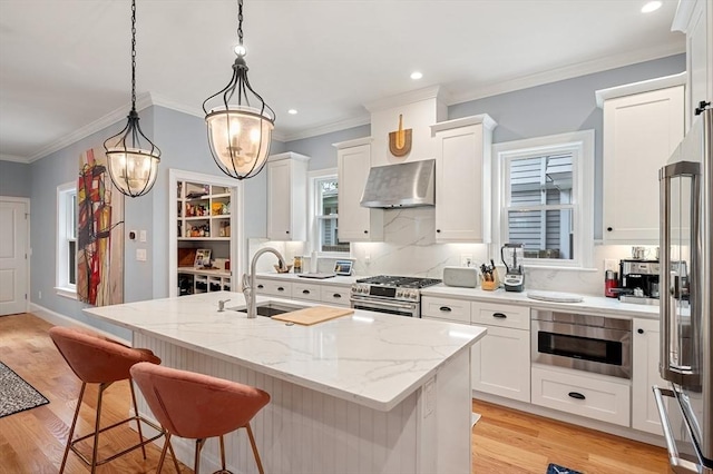 kitchen featuring a center island with sink, light wood-style floors, appliances with stainless steel finishes, wall chimney range hood, and a sink