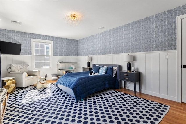 bedroom featuring a wainscoted wall, visible vents, wallpapered walls, and wood finished floors