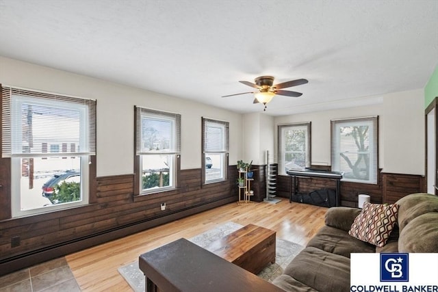 living area with ceiling fan, wooden walls, wood finished floors, baseboard heating, and wainscoting