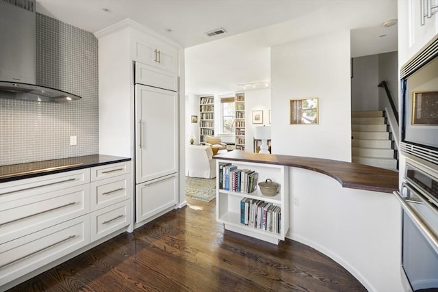 kitchen with dark wood-type flooring, built in appliances, white cabinets, and wall chimney exhaust hood