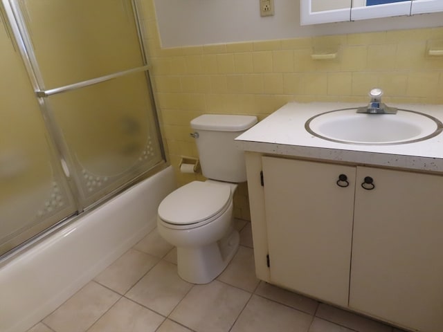 full bathroom with tile walls, tile patterned floors, bath / shower combo with glass door, vanity, and toilet