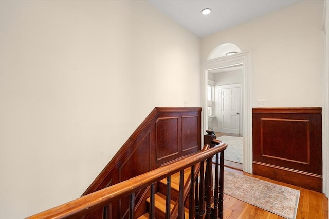 hall with light wood-type flooring, a wainscoted wall, and an upstairs landing