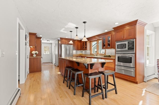 kitchen with light stone counters, a warming drawer, appliances with stainless steel finishes, brown cabinetry, and a baseboard heating unit