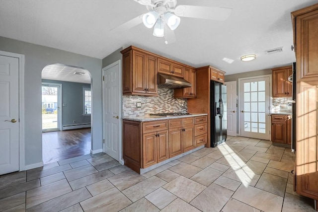 kitchen with black refrigerator with ice dispenser, decorative backsplash, ceiling fan, a baseboard radiator, and stainless steel gas cooktop