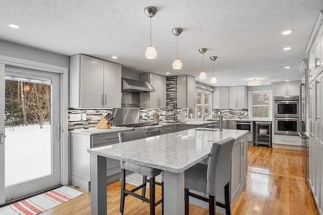 kitchen featuring wall chimney exhaust hood, hanging light fixtures, a center island with sink, gray cabinets, and double oven