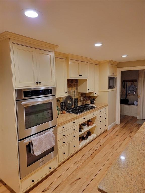 kitchen with cream cabinetry, light stone countertops, light hardwood / wood-style floors, decorative backsplash, and stainless steel appliances