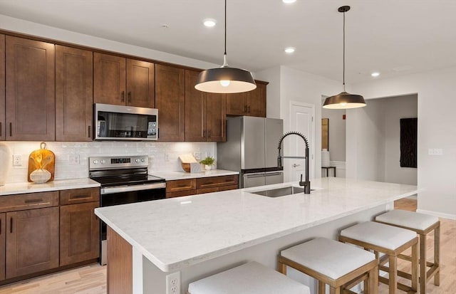 kitchen with a sink, a center island with sink, backsplash, light wood-style flooring, and stainless steel appliances