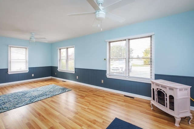 spare room with a ceiling fan, visible vents, a wood stove, wainscoting, and light wood-type flooring