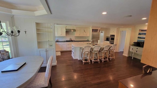 kitchen featuring wall chimney range hood, a kitchen breakfast bar, crown molding, a center island with sink, and dark wood finished floors