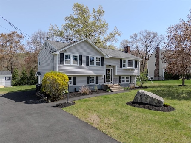 raised ranch with a front lawn, a chimney, a shingled roof, an outbuilding, and a storage shed