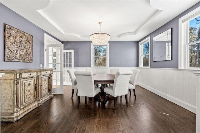 dining area with dark wood-type flooring, a raised ceiling, a wainscoted wall, and an inviting chandelier