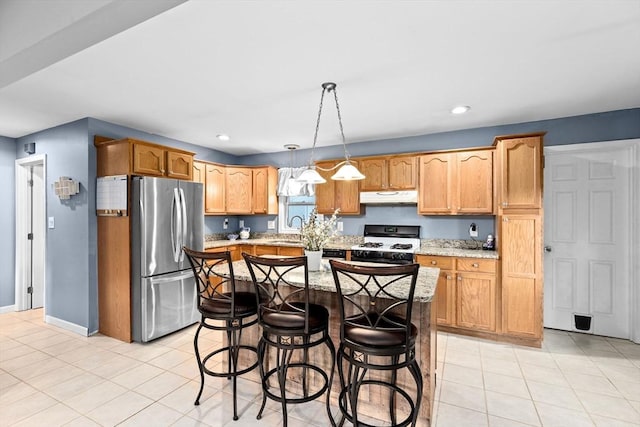 kitchen featuring gas range gas stove, freestanding refrigerator, under cabinet range hood, light stone countertops, and a kitchen breakfast bar
