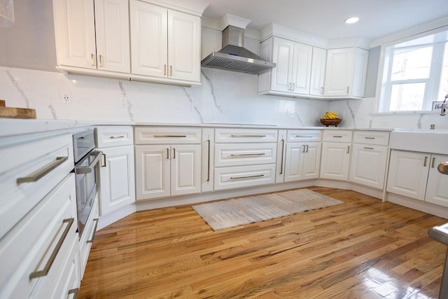 kitchen with wall chimney exhaust hood, light countertops, light wood-style floors, white cabinetry, and backsplash