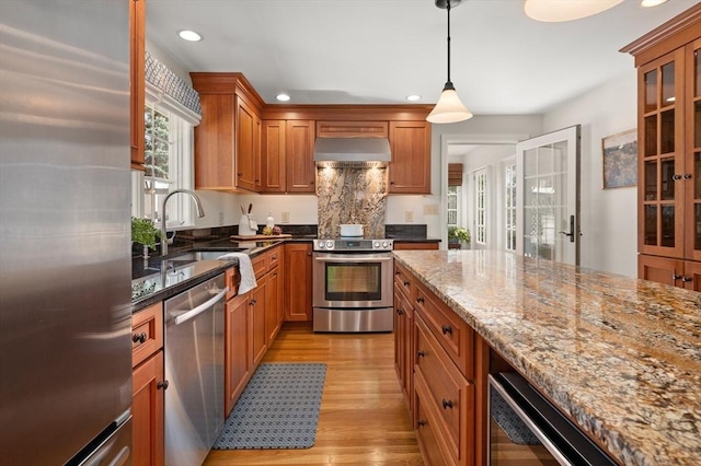 kitchen with dark stone countertops, exhaust hood, brown cabinetry, stainless steel appliances, and a sink