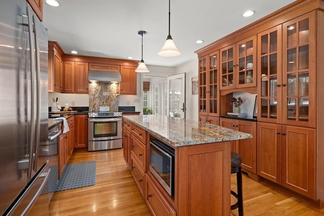 kitchen with light stone counters, brown cabinetry, stainless steel appliances, a kitchen bar, and exhaust hood