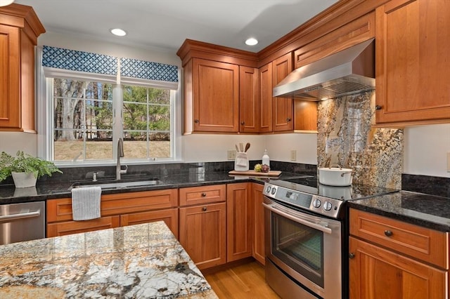 kitchen featuring brown cabinets, a sink, dark stone countertops, appliances with stainless steel finishes, and wall chimney range hood