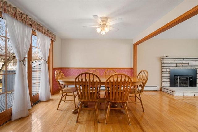 dining room with a wainscoted wall, a fireplace, light wood finished floors, a baseboard heating unit, and a ceiling fan