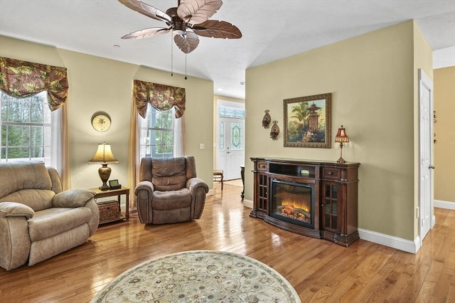 living area with a glass covered fireplace, ceiling fan, baseboards, and hardwood / wood-style flooring