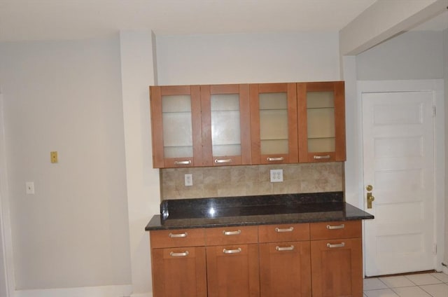 kitchen with light tile patterned floors, tasteful backsplash, brown cabinetry, glass insert cabinets, and dark stone counters