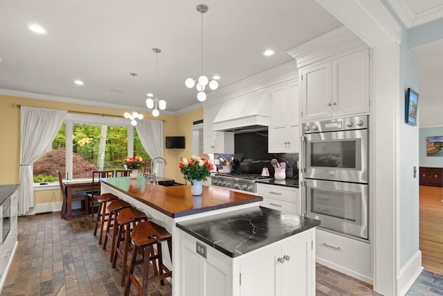 kitchen with stainless steel double oven, dark countertops, crown molding, an island with sink, and custom range hood
