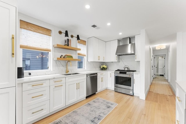 kitchen with stainless steel appliances, wall chimney exhaust hood, a sink, light wood-style floors, and light countertops