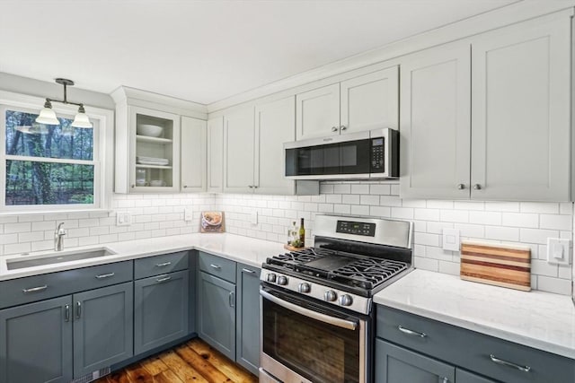 kitchen with gray cabinetry, glass insert cabinets, a sink, stainless steel appliances, and tasteful backsplash