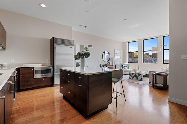kitchen with built in appliances, light wood finished floors, a kitchen breakfast bar, dark brown cabinets, and recessed lighting