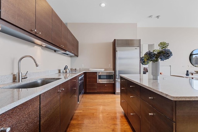 kitchen featuring a sink, built in appliances, light wood finished floors, dark brown cabinetry, and light stone counters