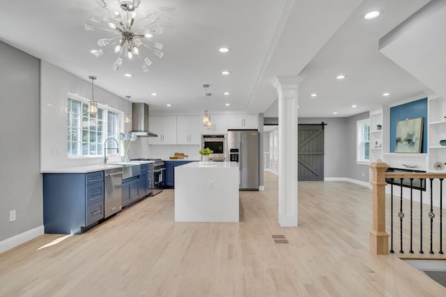 kitchen with blue cabinetry, wall chimney range hood, light countertops, a barn door, and appliances with stainless steel finishes