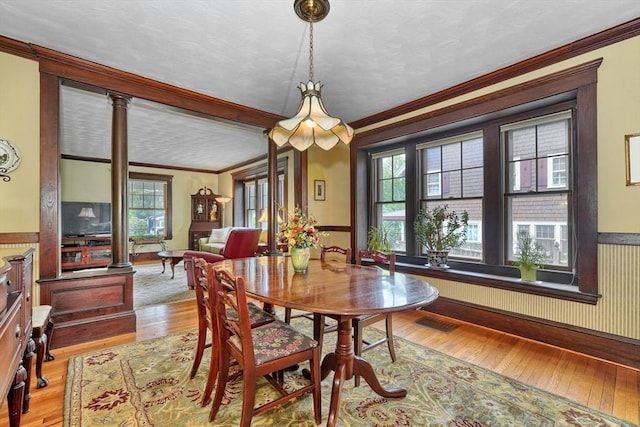dining room with ornamental molding, ornate columns, light wood finished floors, a wainscoted wall, and a textured ceiling