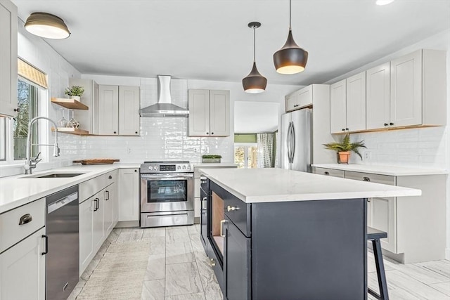 kitchen featuring a kitchen island, hanging light fixtures, stainless steel appliances, wall chimney range hood, and a sink