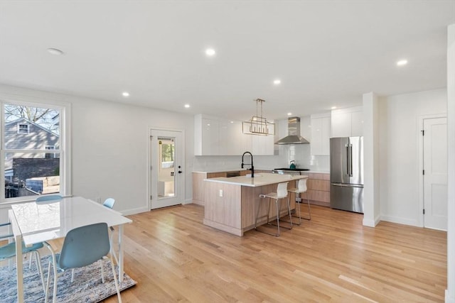 kitchen with wall chimney range hood, a sink, freestanding refrigerator, a center island with sink, and modern cabinets