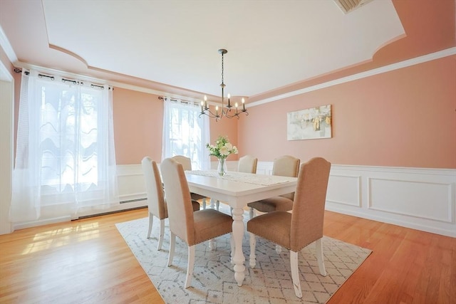 dining room featuring an inviting chandelier, ornamental molding, a decorative wall, a raised ceiling, and light wood-style flooring