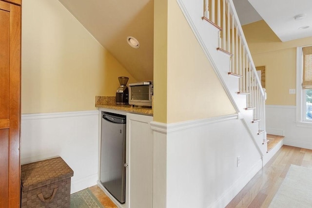 bar featuring light wood-type flooring, a wainscoted wall, stairs, and vaulted ceiling