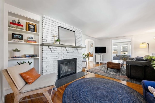 living room featuring built in features, arched walkways, a brick fireplace, and wood finished floors