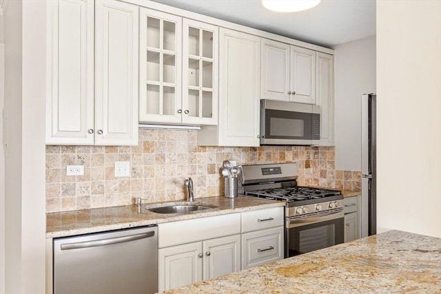 kitchen featuring stainless steel appliances, a sink, decorative backsplash, light stone countertops, and glass insert cabinets