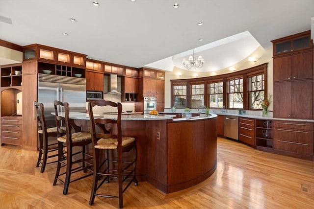kitchen with stainless steel appliances, a large island with sink, light wood-type flooring, a notable chandelier, and wall chimney range hood
