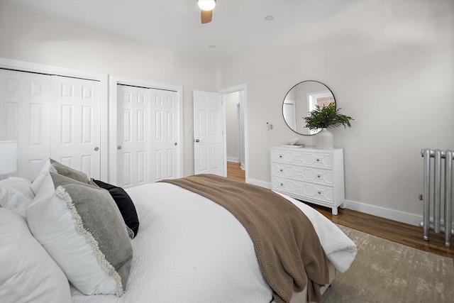 bedroom with radiator, two closets, ceiling fan, baseboards, and wood finished floors