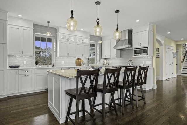 kitchen with wall chimney exhaust hood, white cabinetry, decorative light fixtures, stainless steel microwave, and a kitchen island