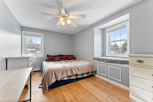 bedroom featuring a baseboard heating unit, multiple windows, ceiling fan, and light wood finished floors