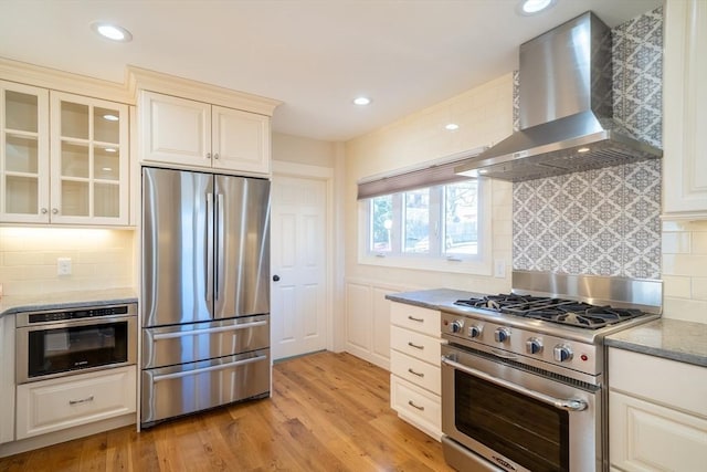 kitchen featuring light wood-style floors, glass insert cabinets, appliances with stainless steel finishes, light stone countertops, and wall chimney range hood