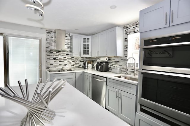 kitchen with stainless steel appliances, light countertops, glass insert cabinets, a sink, and wall chimney exhaust hood