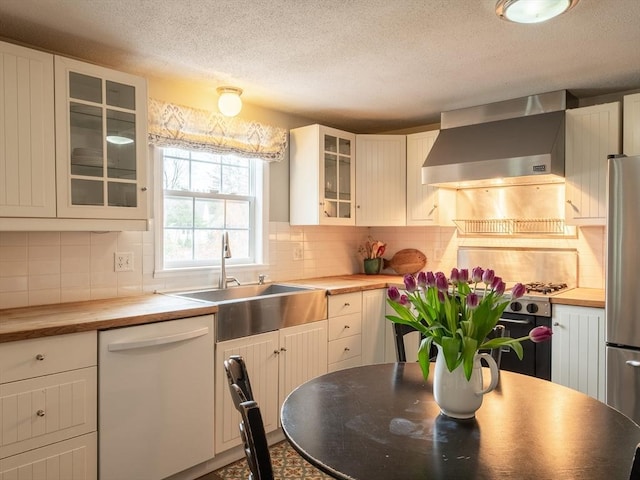 kitchen with dishwasher, butcher block counters, a sink, wall chimney range hood, and freestanding refrigerator